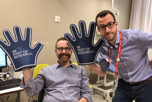 Two men smiling and holding up Be Part of Research foam rubber hands.